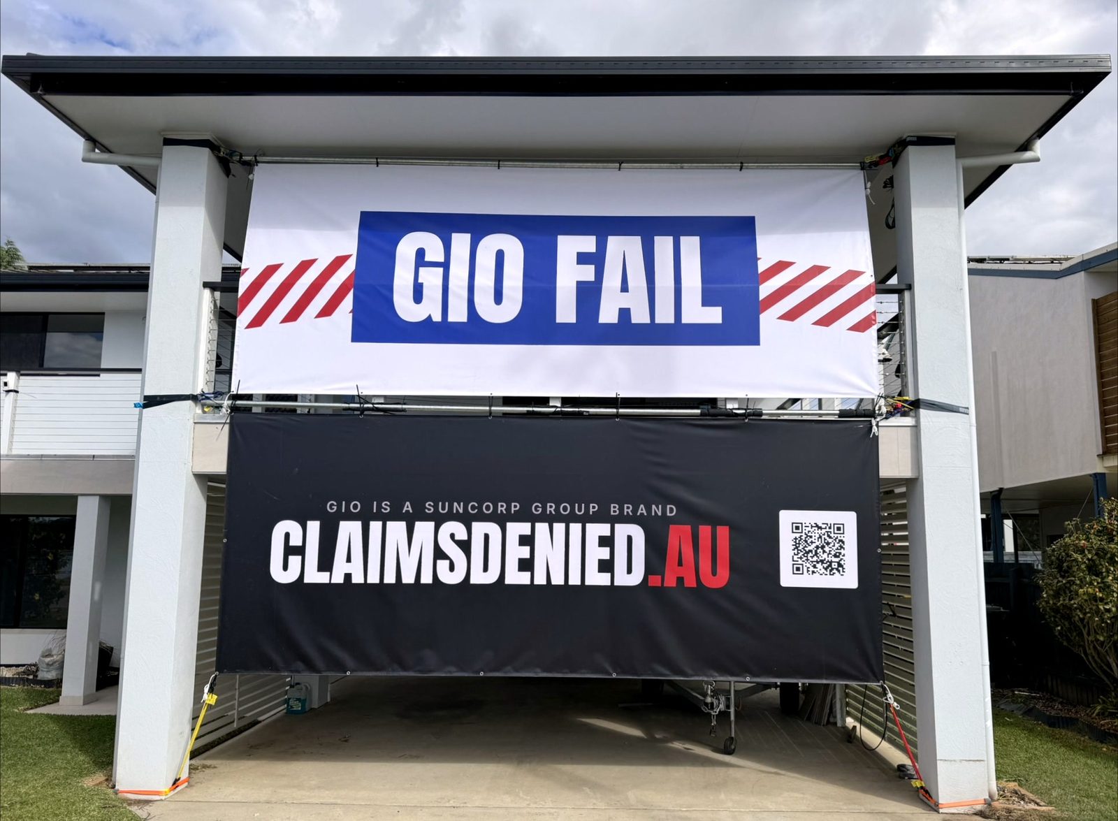 Two large banners mounted on the front of the house: GIO FAIL on a blue block over torn red-and-white barrier tape, and below it a black banner reading GIO IS A SUNCORP GROUP BRAND, CLAIMSDENIED.AU, with a QR code.
